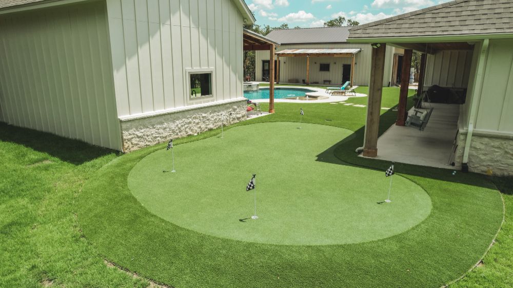 An aerial view of a custom artificial turf putting green with four flags in a luxurious backyard, featuring white buildings with stone bases and a swimming pool in the background, designed for a resort-style outdoor living space.
