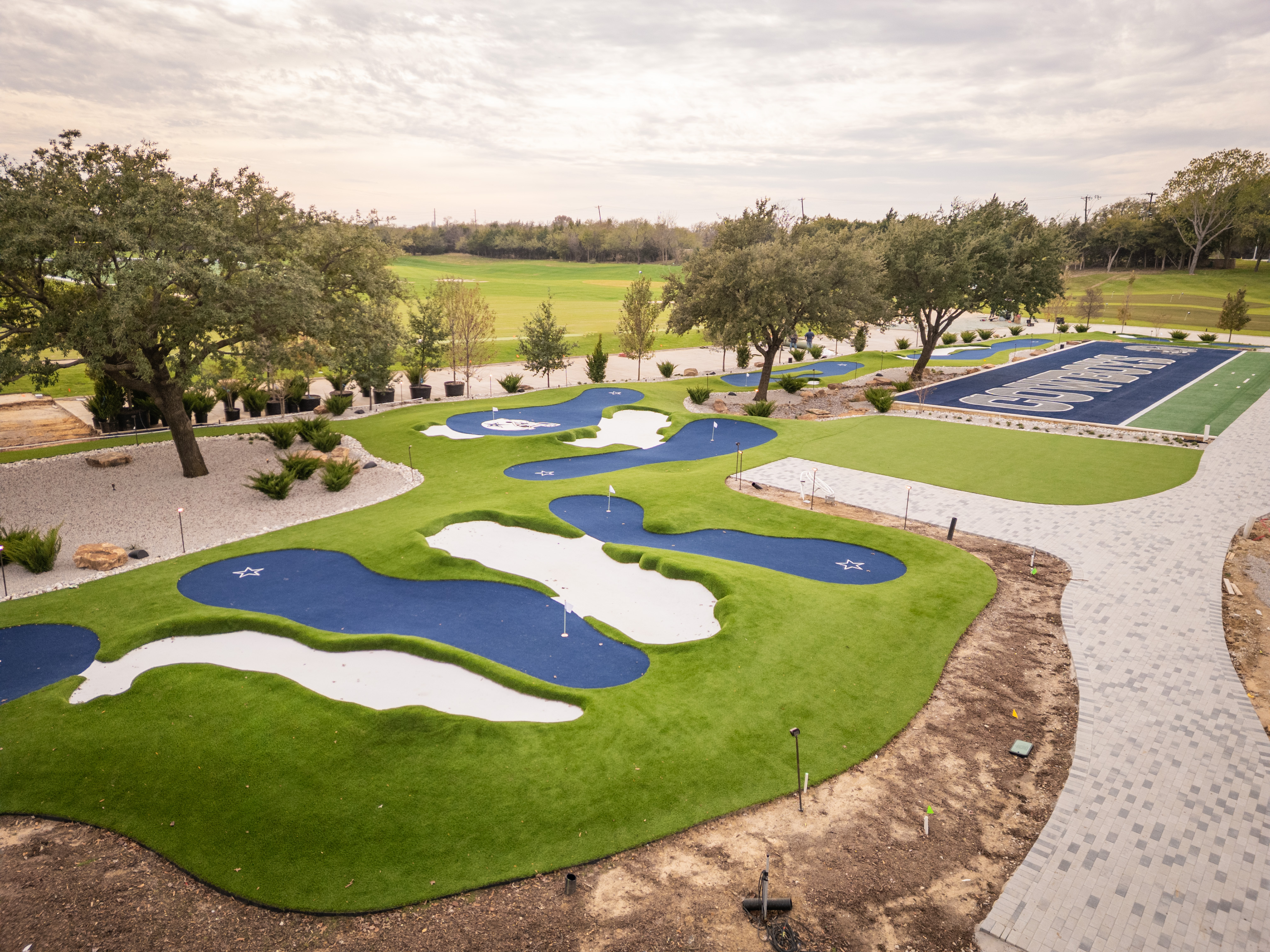 Aerial view of the Cowboys Golf Club's new 18-hole artificial turf putt-putt course, featuring blue and white themed greens, surrounded by landscaping and part of the football field.