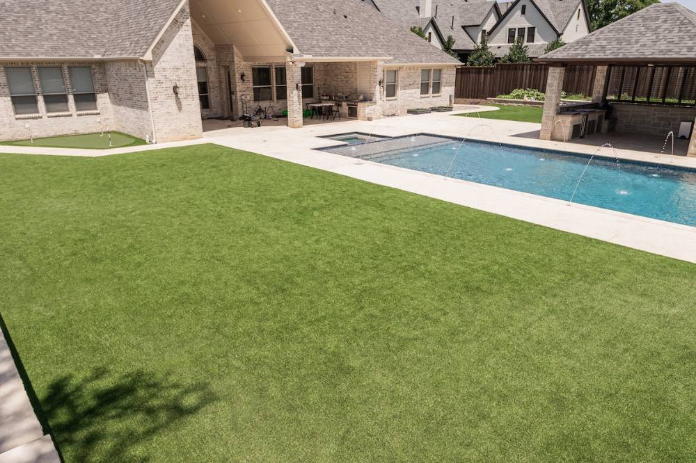 View of a luxurious backyard featuring a large artificial turf lawn, a sparkling swimming pool with fountains, a stone patio, an outdoor kitchen under a pergola, and a small putting green, all complementing a modern brick home.