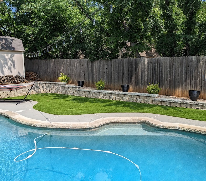 Residential backyard with artificial turf installed around a swimming pool, alongside a retaining wall, enhancing the landscape with low-maintenance synthetic grass.