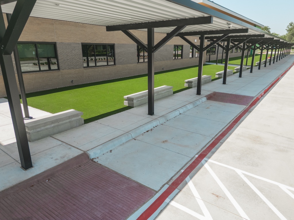 Installation of durable synthetic grass in a low-maintenance school grounds walkway area. This school artificial turf solution enhances enhanced curb appeal and eliminates soil erosion beneath covered structures, providing a clean, bright, and vibrant accent that requires minimal upkeep for the school and its staff