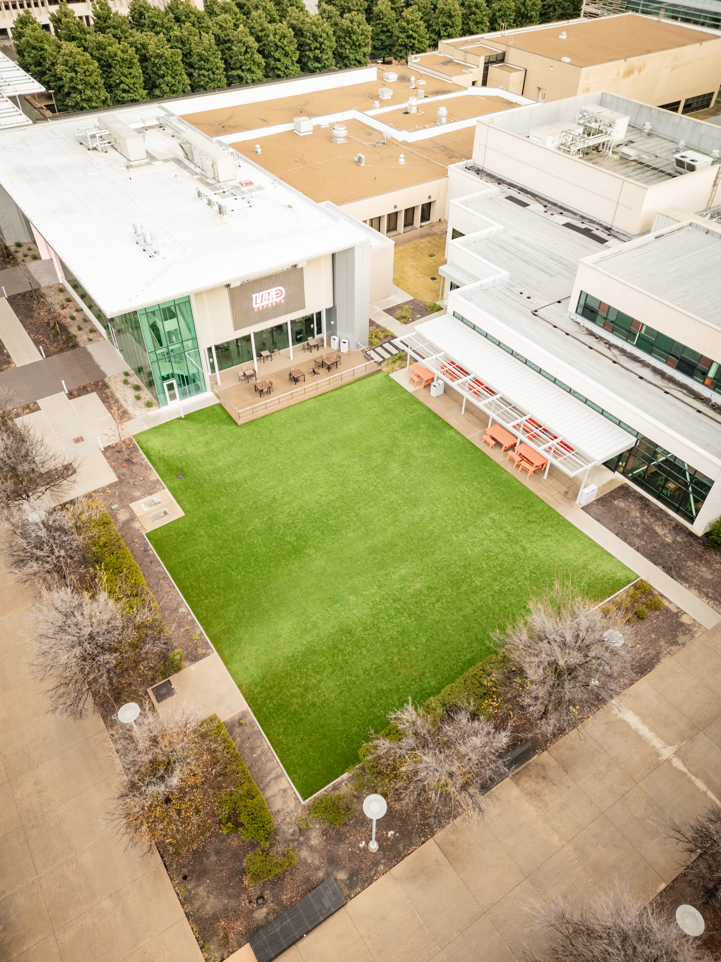 Aerial view of a university campus courtyard featuring newly installed artificial turf, surrounded by modern buildings, walkways, and outdoor seating areas, designed for student engagement and events.