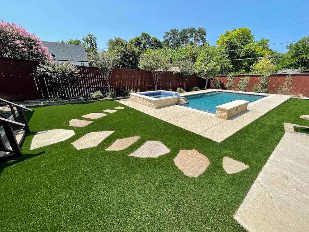 Detail of synthetic grass installation around a pool and jacuzzi area. Custom-cut flagstone pavers create a unique path across the durable, low-maintenance artificial turf. Ideal backyard landscaping solution.