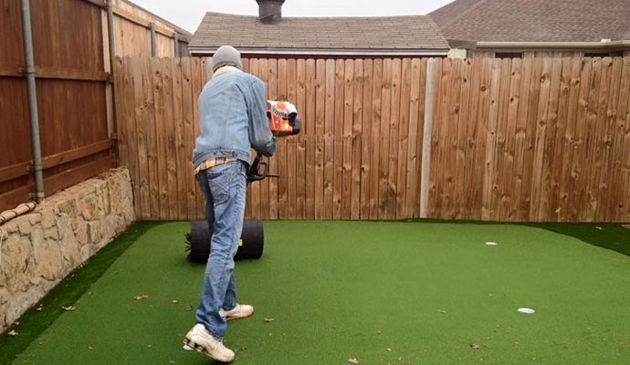 Worker using a power broom on an artificial turf putting green.