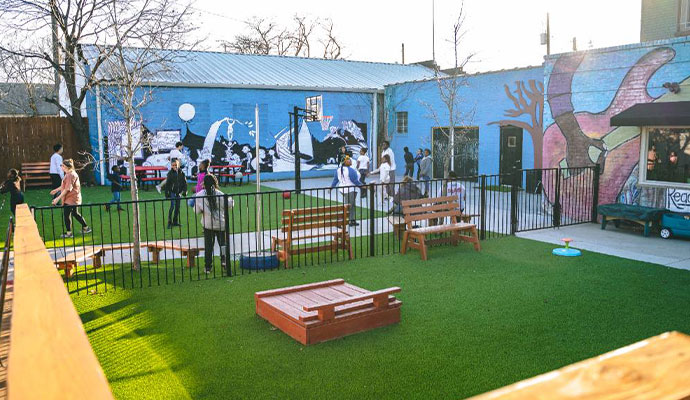 Children and students gathered in an outdoor school courtyard with artificial turf and benches